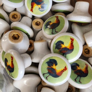 May include: A collection of white ceramic knobs with rooster designs. The knobs have a green background and feature roosters in various poses.