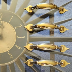 May include: A close-up shot of a clock face and gold-coloured handles. The clock has a dark grey face with gold numerals and hands. The handles are oval-shaped with decorative ends. The word "Elgin" is visible on the clock face.