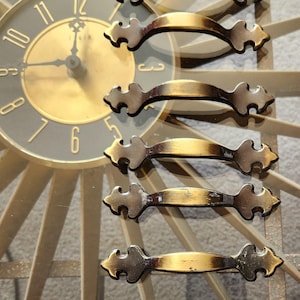 May include: Eight gold-toned cabinet door pulls with ornate, decorative details. The pulls have a slightly aged, antique look.