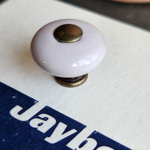 May include: A light purple cabinet knob with a brass-colored base and top. The knob is sitting on a white surface with the text "Jaybee Ajax" in a blue rectangle.