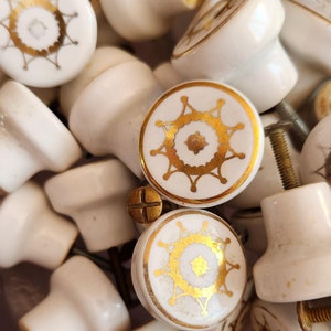 May include: A collection of white ceramic cabinet knobs with gold accents. The knobs feature a variety of geometric designs.