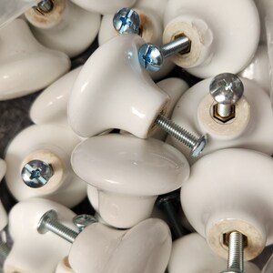 May include: A close-up of a pile of white ceramic cabinet knobs with silver screws. The knobs are round and have a smooth, glossy finish.