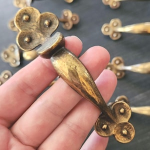 May include: Antique brass cabinet pulls with a clover-shaped design. The pulls have a curved handle and decorative clover-shaped ends. The brass has a warm, aged patina. Several pulls are visible, suggesting a set for furniture restoration or new builds.