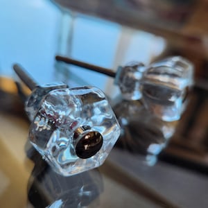May include: Two clear glass cabinet knobs with a faceted hexagonal shape. Each knob features a central silver-toned metal screw attachment at the front, with a long metal bolt extending from the back for installation on furniture or doors.