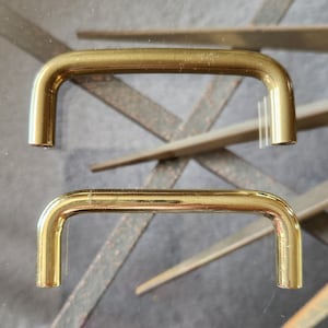 May include: Two gold metal cabinet door pulls. The pulls are shaped like a D with rounded edges. The pulls are about 3 inches long.