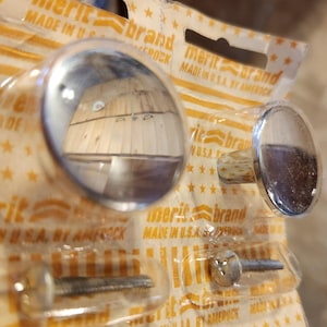 May include: Two silver-toned cabinet knobs with screws. The knobs are round and have a reflective surface. The packaging is yellow with the text "merit brand made in USA by Amerock".