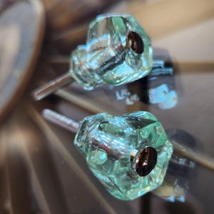 May include: Two light green, translucent glass cabinet knobs with a faceted, geometric shape. Each knob features a central dark metal circular insert and a silver-toned metal bolt extending from the back for mounting purposes.