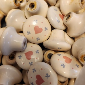 May include: A collection of off-white ceramic cabinet knobs. Some knobs feature a pink heart design with small blue floral accents. The knobs have a variety of shapes, including round and heart-shaped, and are piled together.