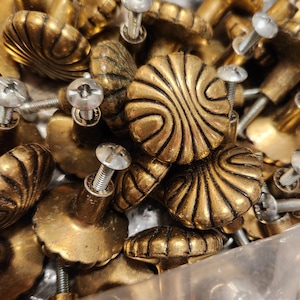 May include: A close-up of a pile of antique gold-toned cabinet knobs with intricate designs. The knobs have a variety of shapes and sizes, and some have a decorative screw head.