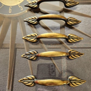 Peut inclure: Cinq poignées de tiroir de cabinet dorées avec des extrémités en forme de feuille. Les poignées sont disposées en rangée, les extrémités des feuilles pointant dans des directions opposées.