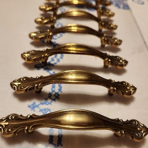 May include: A set of ten antique brass cabinet door pulls with ornate designs. The pulls are arranged in a row, with the largest pull at the top and the smallest pull at the bottom.