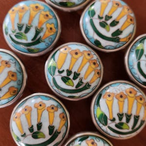 May include: A collection of round cabinet knobs featuring a light blue background with a hand-painted floral pattern of yellow trumpet-shaped flowers and green leaves, each enclosed by a thin silver-toned metal rim and a smooth glossy finish.