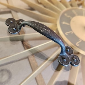 May include: Black metal cabinet door handle with a clover-shaped design. The handle has a distressed finish and is suitable for a rustic or farmhouse style.