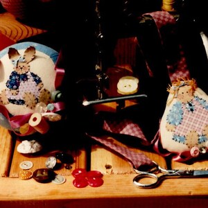 May include: A vintage sewing pattern for dolls, titled "Dolls by Ruth" and "Sew Helpful RC #130". The pattern features four dolls with different designs and colors, including a blue and white doll, a pink and white doll, a blue and pink doll, and a pink and white doll. The dolls are surrounded by sewing supplies, including scissors, buttons, and thread.