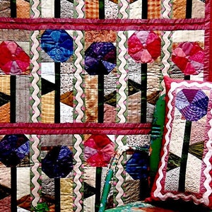 May include: A quilt pattern titled "#175 Petunia Patch" from the "Joined at the Hip" collection. The quilt features a colourful floral design with a red, green, and blue colour scheme. The quilt is displayed on a chair with a floral cushion.
