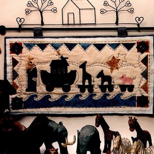May include: A colourful quilt depicting Noah's Ark with animals on a blue sea. The quilt is hanging on a wall with a wooden house and two trees above it. There are wooden animal figurines in front of the quilt, including a zebra, giraffe, elephant, and a bull. The text "Noah's Rolling Ark" is at the top of the image, and the text "From the Powder Mill" is at the bottom.