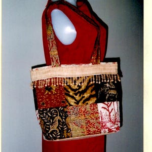 May include: A red quilted purse with a patchwork design. The purse has a long strap and a fringe trim. The purse is on a mannequin.