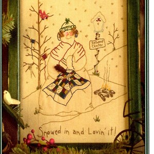 May include: A cross-stitch embroidery design featuring a snowman sitting by a fire with a quilt. The design is framed in a green frame and includes the text "Snowed in and Lovin' it!" and "Retweet Center".