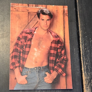 May include: A man wearing a red and black plaid shirt with the top unbuttoned, revealing his bare chest. He is wearing blue jeans and has a serious expression on his face.
