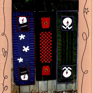 May include: Three fabric wall hangings with different designs. The leftmost hanging is blue with white snowflakes and a snowman design. The middle hanging is black and red with a checkered pattern and a red apple design. The rightmost hanging is green with a white bunny design.