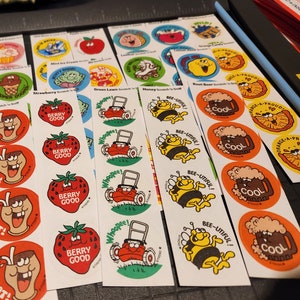 May include: A collection of vintage scratch and sniff stickers featuring various designs. Stickers include images of strawberries with "Berry Good" text, a lawnmower with "Whoopee!", and a bee with "Bee-utiful!". Other stickers depict ice cream, root beer, and orange slices.