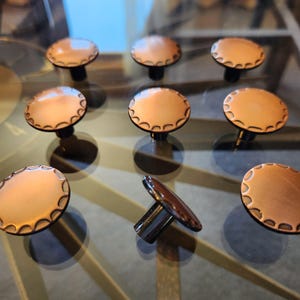 May include: A collection of nine copper-colored cabinet knobs with a scalloped edge design. The knobs have a round, flat top and a dark, cylindrical base. The knobs are arranged on a reflective surface.