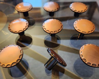 May include: A collection of nine copper-colored cabinet knobs with a scalloped edge design. The knobs have a round, flat top and a dark, cylindrical base. The knobs are arranged on a reflective surface.