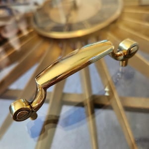 May include: A close-up of a gold-colored furniture handle. The handle has a curved design with rounded ends and a polished finish. The background shows a gold-colored, sunburst-style design.