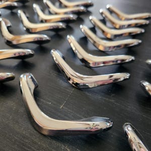 Peut inclure: Plusieurs poignées de porte chromées brillantes, disposées sur une surface sombre. Chaque poignée présente une forme incurvée et ergonomique, reflétant la lumière. L'ensemble dégage une impression de modernité et d'aspect industriel.