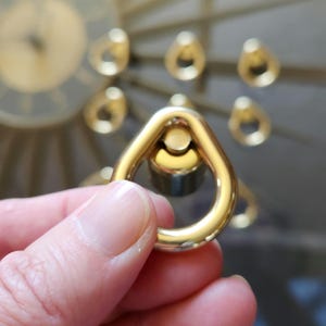 May include: A close-up of a gold-colored, triangular-shaped metal object with a circular opening. The object is held in a person's hand. Other gold-colored, teardrop-shaped objects are in the background, out of focus.