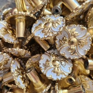 May include: Close-up of a pile of ornate gold-colored cabinet knobs. Each knob features a white floral design with gold accents. The knobs have a classic, vintage aesthetic, suitable for furniture restoration or decorative projects.