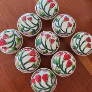 May include: A collection of eight round ceramic cabinet knobs, each featuring a white background with a hand-painted design of red tulip-like flowers on green stems and leaves. The knobs have a smooth, glossy finish and a silver-toned metal base.