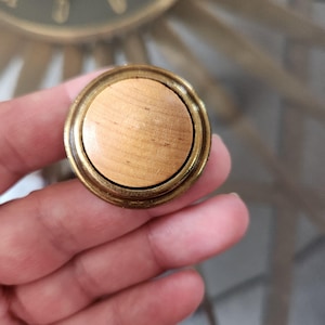 May include: A circular wooden knob with a light brown, wood-grain centre. The knob is framed by a brass-coloured metal rim. This is likely a furniture hardware component, such as a drawer pull or cabinet knob.