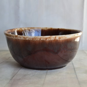 May include: A large, round, brown ceramic mixing bowl with a glossy finish. The bowl has a wide rim with a lighter brown and white edge. The interior of the bowl reflects the light. The bowl is sitting on a light-colored surface.