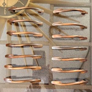 May include: A set of 12 antique-style cabinet pulls in a brushed copper finish. The pulls are all the same size and shape, with a curved design. They are arranged in two rows of six, with each row offset from the other.
