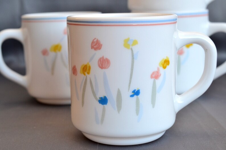May include: Two white ceramic mugs with a floral design. The mugs have a blue, pink, and yellow floral pattern. The mugs are on a gray surface.