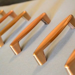 May include: Five brushed copper cabinet pulls with a simple, modern design. The pulls are arranged in a row, with the top pull slightly offset from the others.