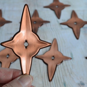 May include: A set of ten copper-coloured star-shaped metal pieces with a hole in the centre. The stars are arranged in a scattered pattern on a light wood surface.