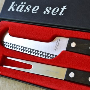 May include: A black box with the words "käse set" on the lid. The box is open and contains two silver cheese knives with black handles. The knives have white dots on the handles.