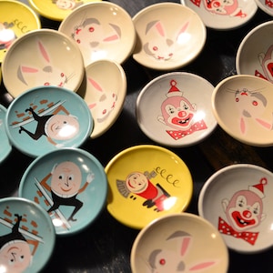 May include: A collection of small, colourful ceramic plates with various whimsical designs. The plates feature illustrations of clowns, bunnies, and other characters.