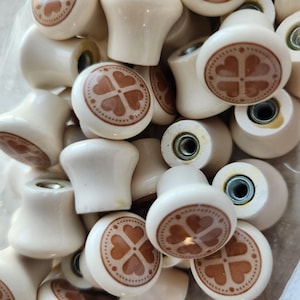 May include: A collection of off-white ceramic cabinet knobs. Each knob features a brown clover design on the front. The knobs have a rounded shape with a metal screw for attachment. The knobs are suitable for furniture restoration or DIY projects.