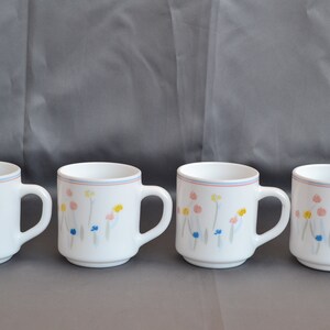 May include: Four white ceramic mugs with a floral design in shades of pink, yellow, blue, and gray. The mugs have a blue and red rim.