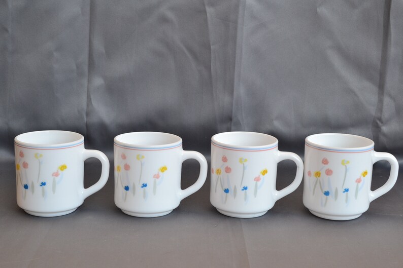 May include: Four white ceramic mugs with a floral design in shades of pink, yellow, blue, and gray. The mugs have a blue and red rim.
