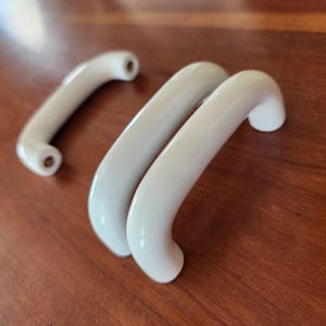 May include: Three white ceramic cabinet pulls with a smooth, arched, semi-circular design. Each handle features a glossy finish and two circular mounting points with visible brass-colored metal inserts for screw attachment at the ends.