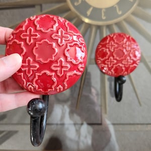 May include: Two red ceramic wall hooks with a decorative pattern. The hooks have a glossy finish and a black metal hook. The design features a repeating pattern of crosses and floral shapes.