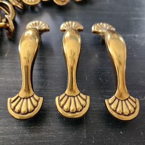 May include: Close-up of several antique brass drawer pulls. The pulls have a curved, elongated design with a fan-shaped base and a decorative top. The brass has a warm, aged patina, suggesting vintage or retro style. The pulls are arranged on a dark surface.