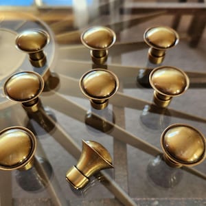 May include: A collection of nine antique brass cabinet knobs and one decorative brass horn-shaped pull. The knobs have a rounded, mushroom-like design. The hardware is displayed on a reflective surface.