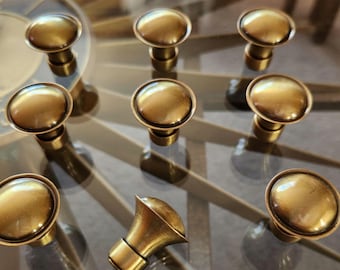 May include: A collection of nine antique brass cabinet knobs and one decorative brass horn-shaped pull. The knobs have a rounded, mushroom-like design. The hardware is displayed on a reflective surface.