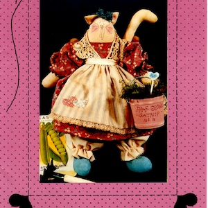 May include: A handmade catnip doll dressed in a red and white floral dress with a white apron and a blue bow. The doll is holding a small bucket with the text "Home Grown Catnip 10¢ 1lb". The doll is 12 inches tall.