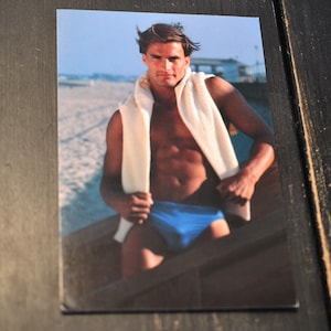 May include: A man wearing blue swim trunks and a white towel around his neck stands on a wooden boat. He is on a beach with sand and water in the background.